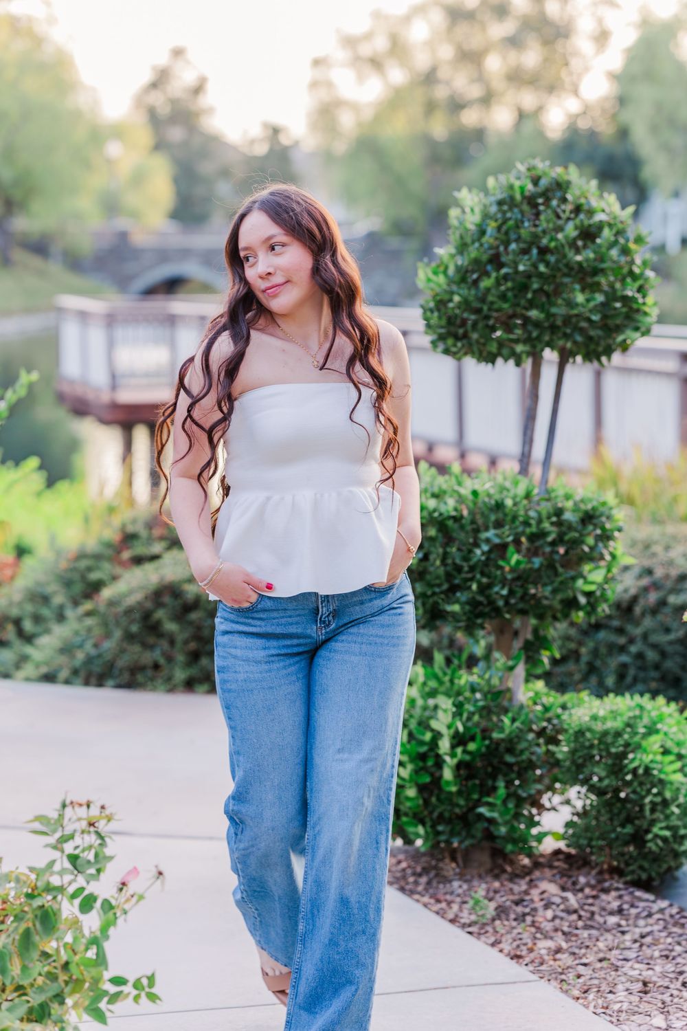 A woman in jeans and a white top stands on a landscaped path with manicured shrubs and a pond view.