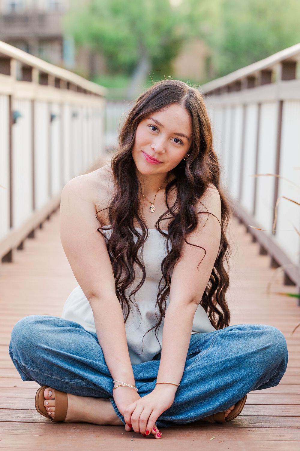 Woman in casual outfit sits cross-legged on wooden deck with long wavy hair and natural smile.