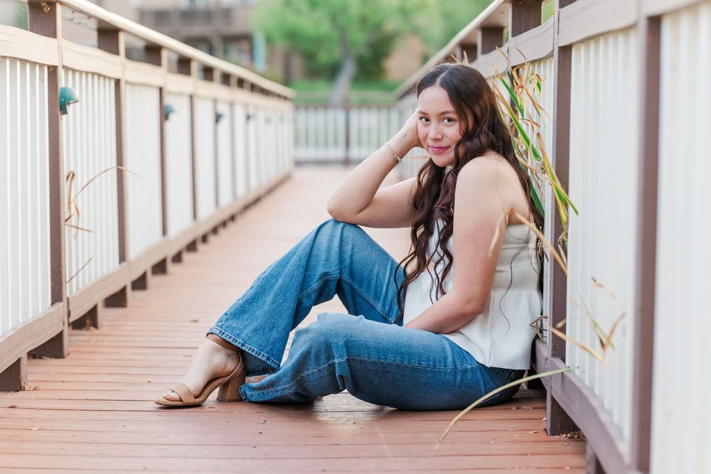 A casual portrait of someone sitting on a wooden boardwalk wearing jeans and brown heels on a sunny day.