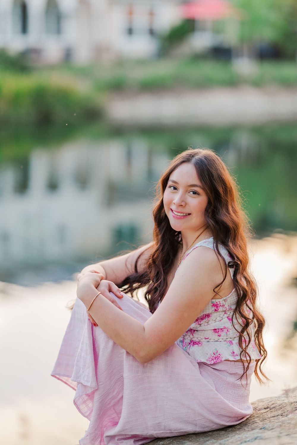 A woman in a pink floral dress sits by a reflective pond during sunset with buildings visible in the background.