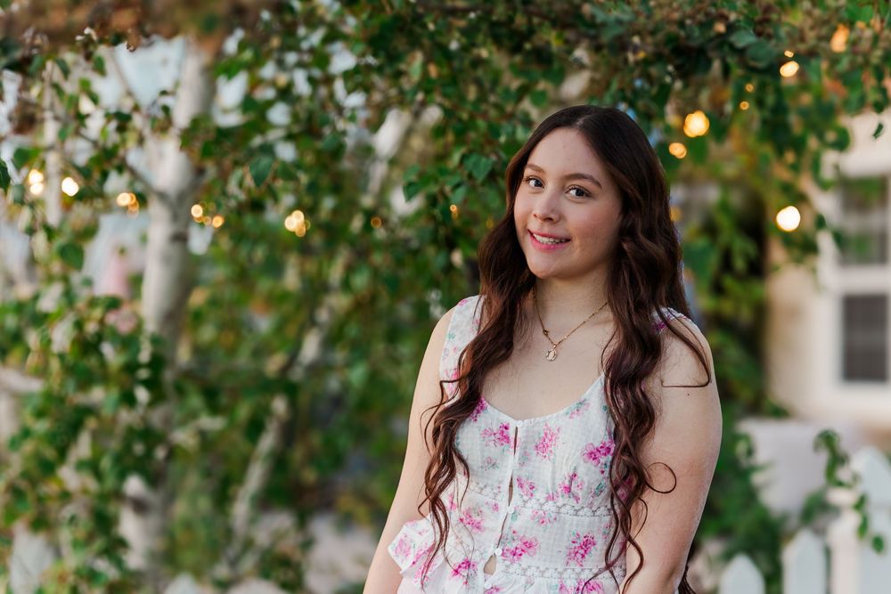 A woman in a floral dress poses among green foliage with soft natural lighting filtering through the trees.