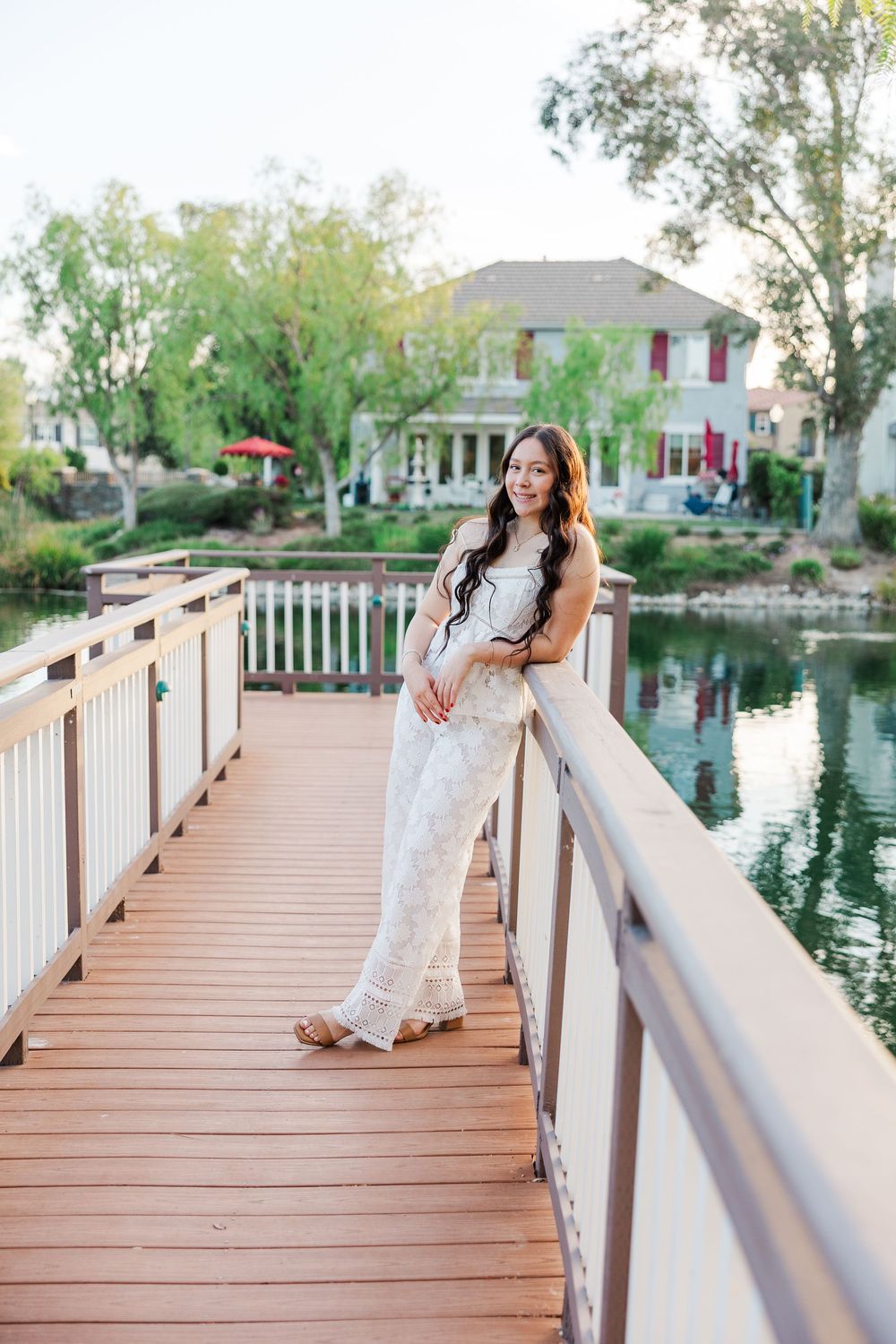 A woman in a white lace dress poses on a wooden bridge overlooking a scenic pond and landscaped grounds at sunset.