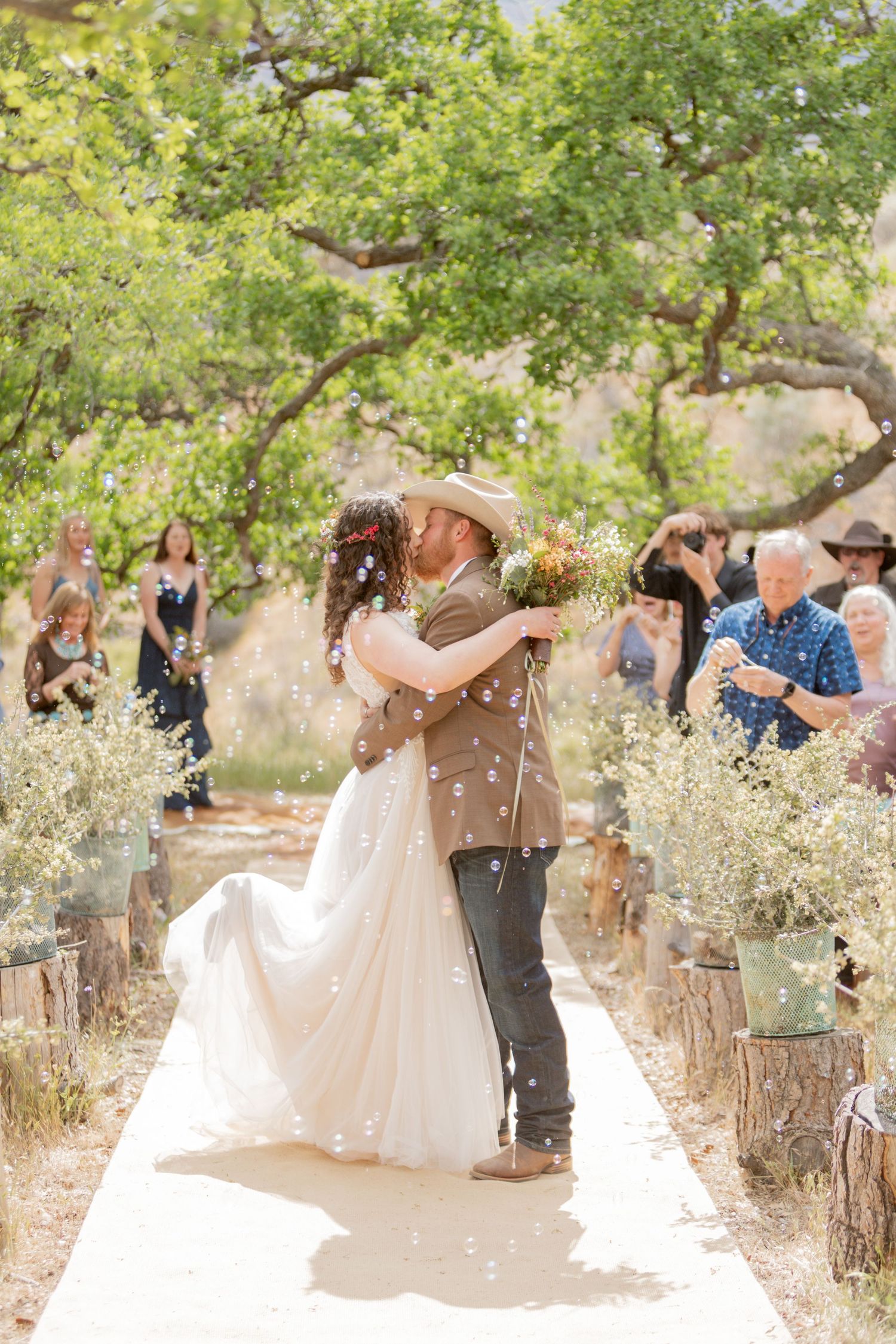 A joyful outdoor wedding celebration with guests throwing confetti under green trees in a natural setting.