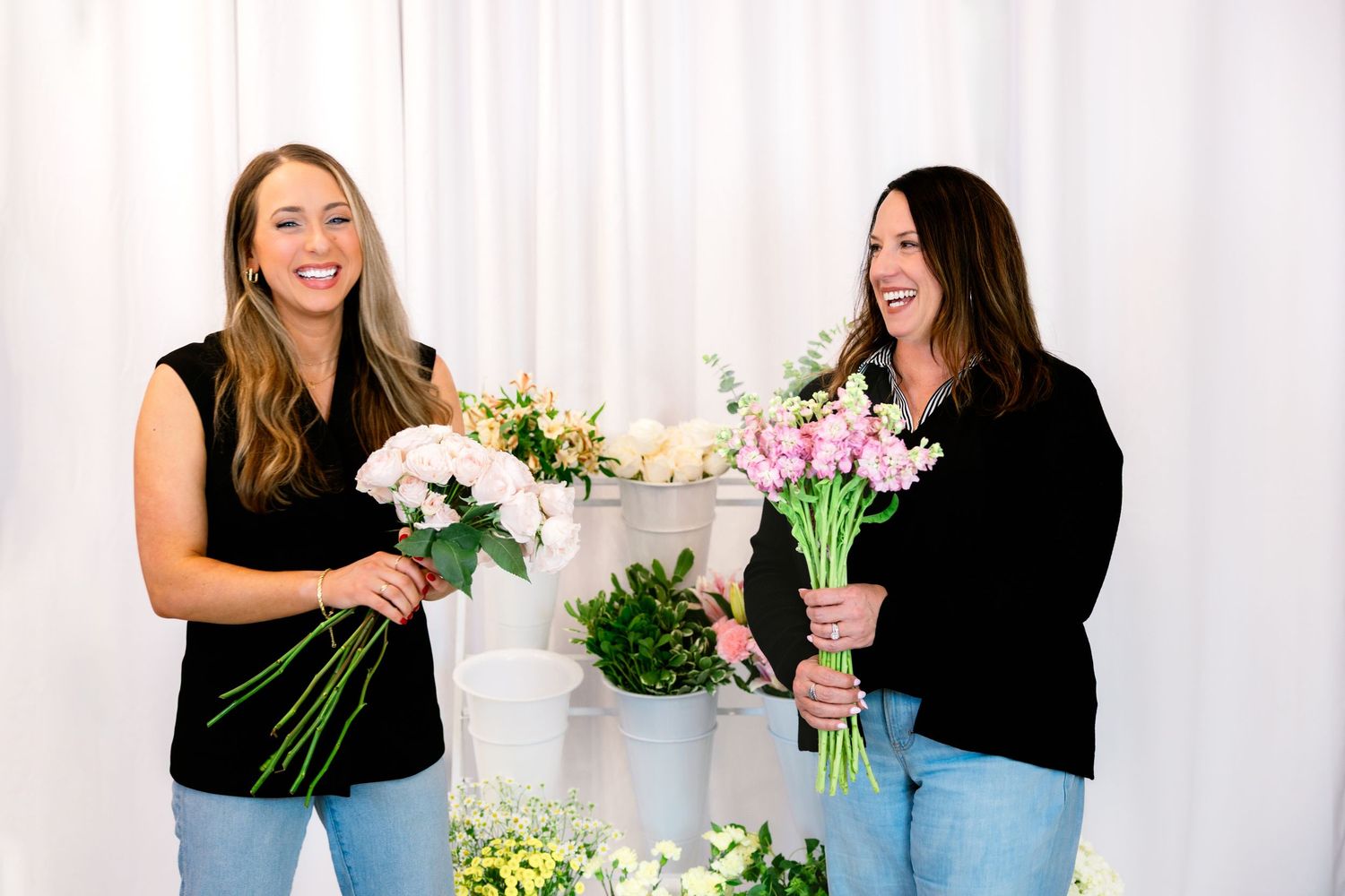 Two people arrange fresh white flowers and greenery for floral design while laughing together in a bright studio space.