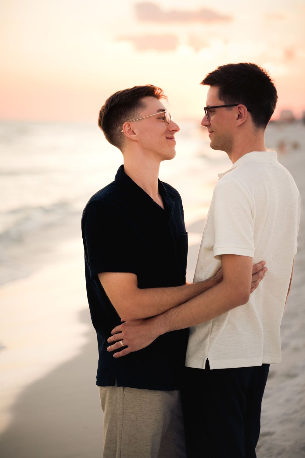 A romantic couple embraces on a beach at sunset with soft pink skies in the background.