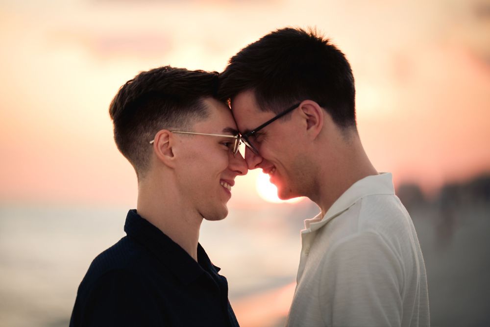 A couple stands close together in silhouette against a warm sunset sky with soft orange and pink tones.