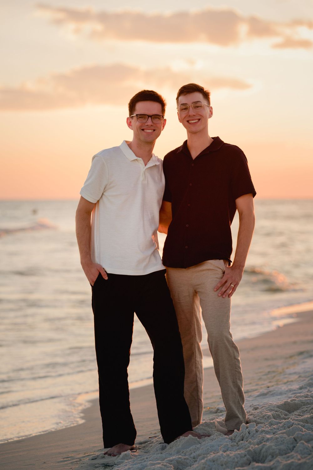 A couple poses together on a sandy beach at sunset with the ocean waves in the background.