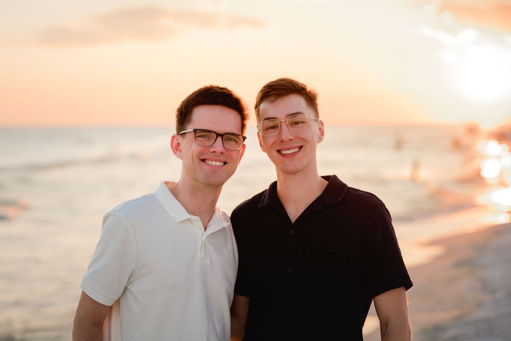 Two people smile and embrace on a beach at sunset with warm orange sky in the background.