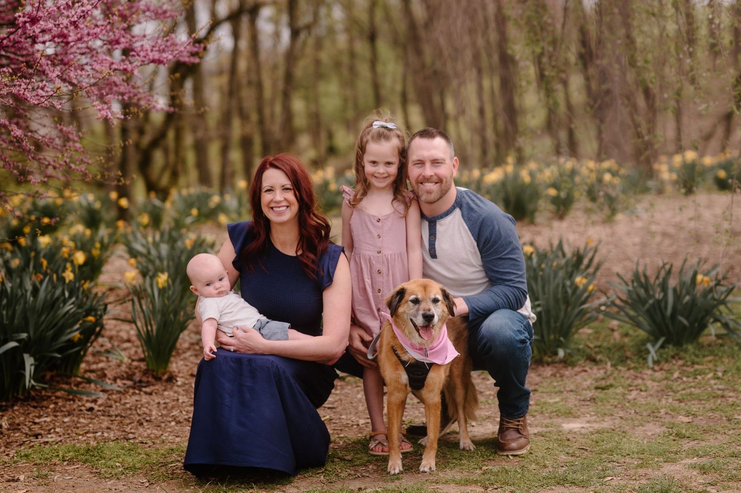 A family poses together with their golden retriever dog in a wooded outdoor setting with spring blooms.