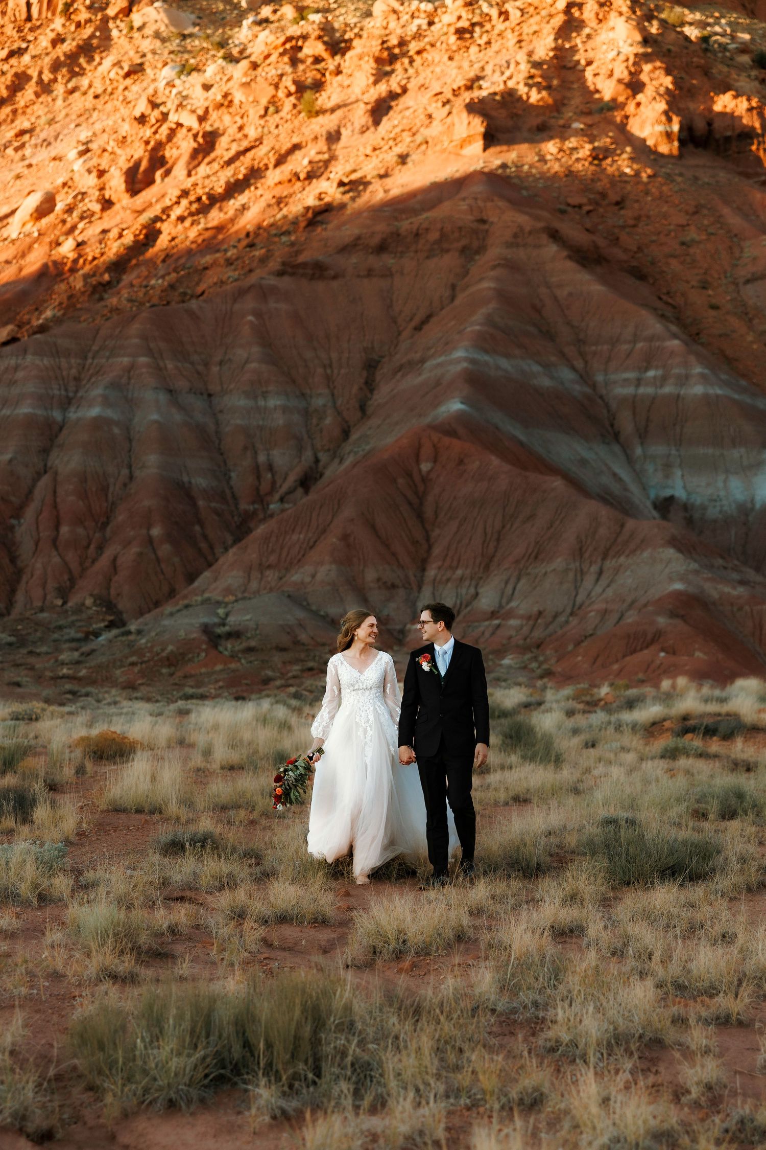 Bride and groom walking hand in hand through a stunning red rock New Mexico desert landscape at sunset.