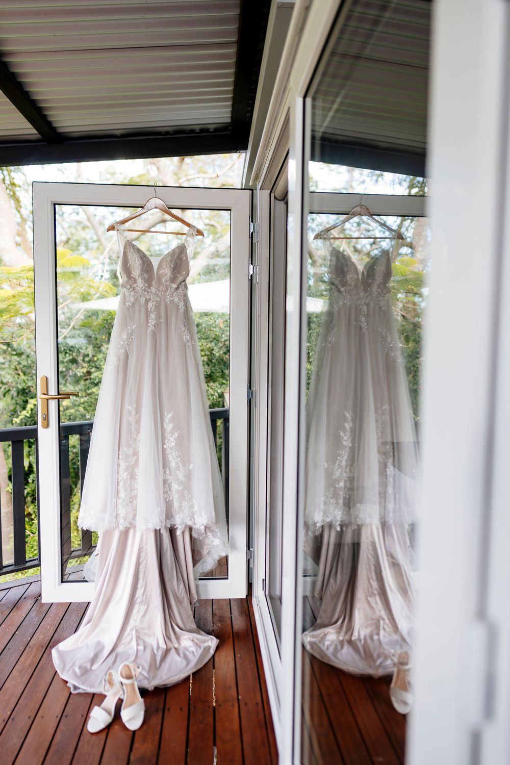 A white wedding dress hangs on display in a sunlit room with wooden deck flooring and glass windows.