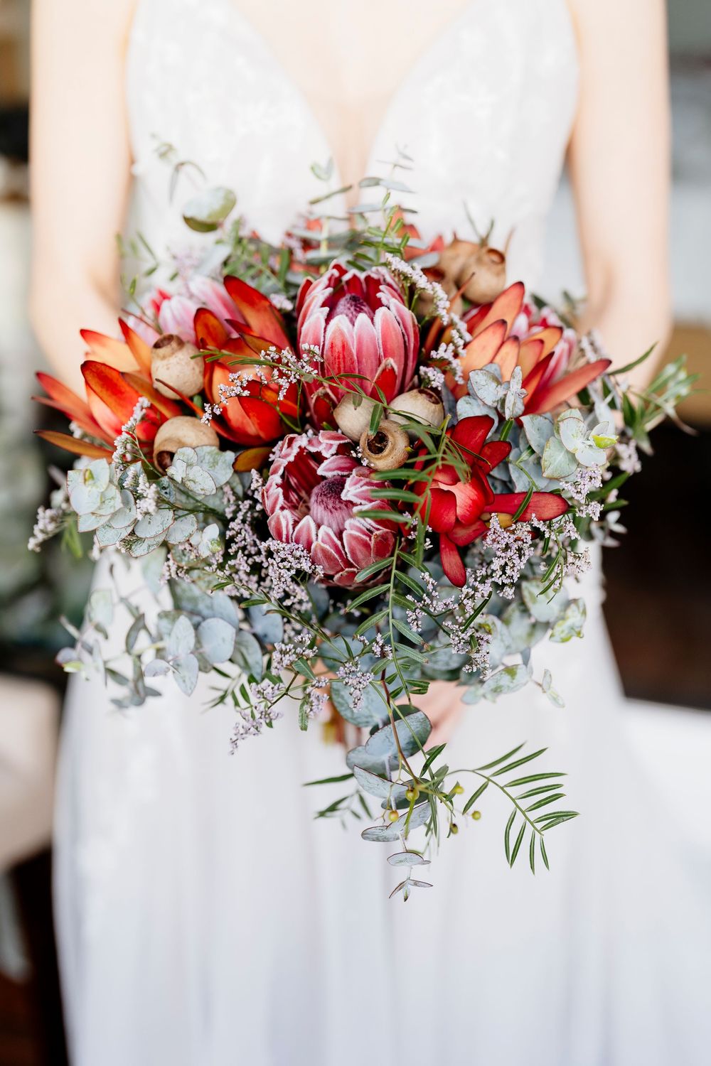 A row of elegant bridal bouquets featuring red protea flowers, white blooms and silvery eucalyptus leaves against a light background.