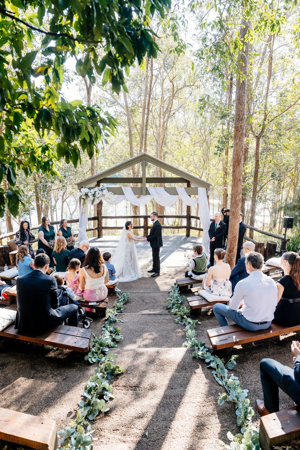 Outdoor wedding ceremony under a rustic wooden pavilion with guests seated on wooden benches in a wooded setting.