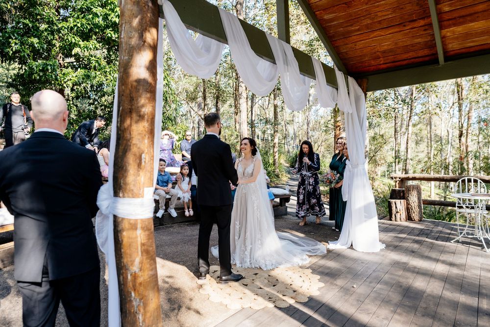 A wedding ceremony takes place under a rustic wooden pavilion decorated with flowing white fabric in an outdoor forest setting.