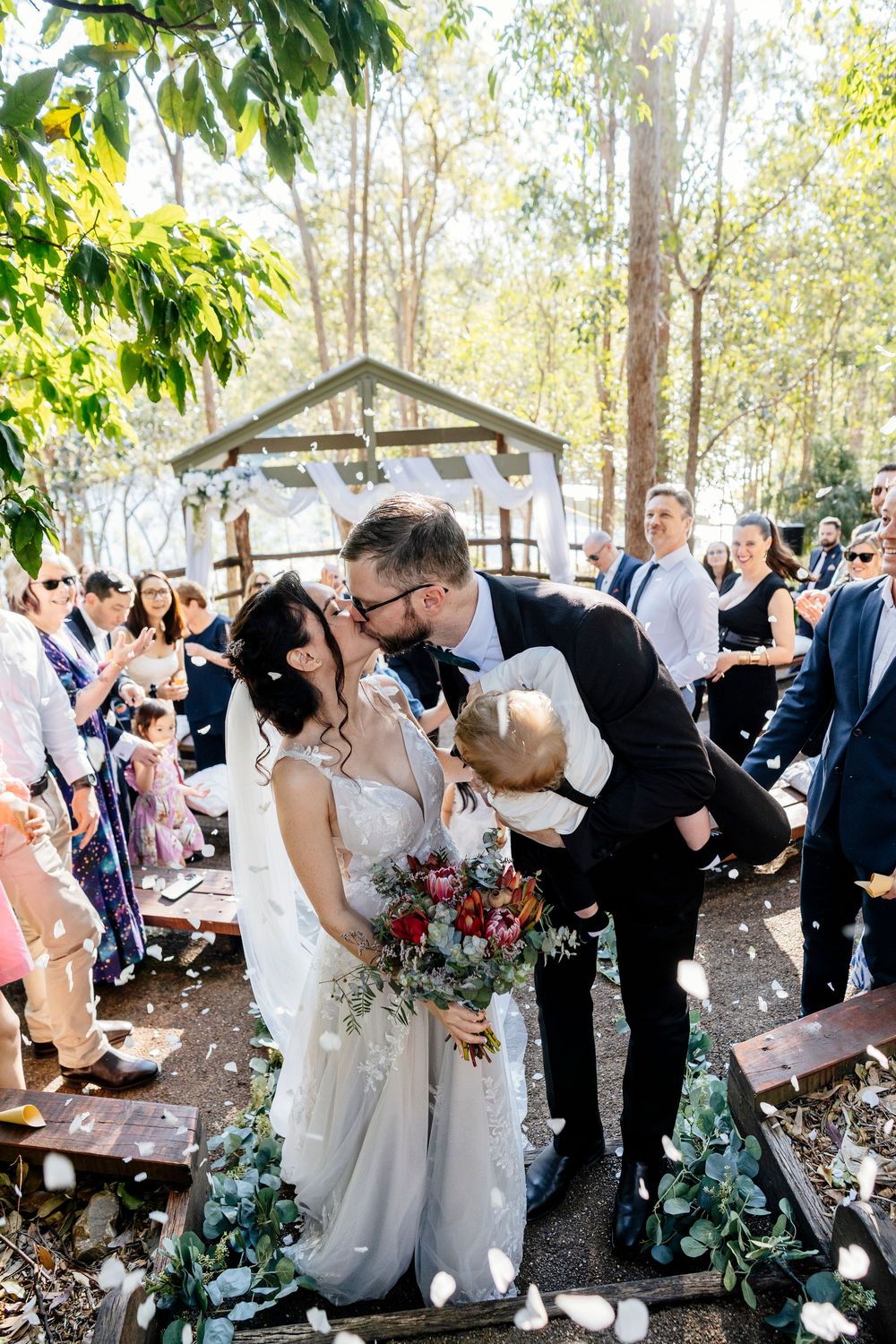 A romantic outdoor wedding celebration with guests watching couples share a kiss under tree canopy in a natural setting.