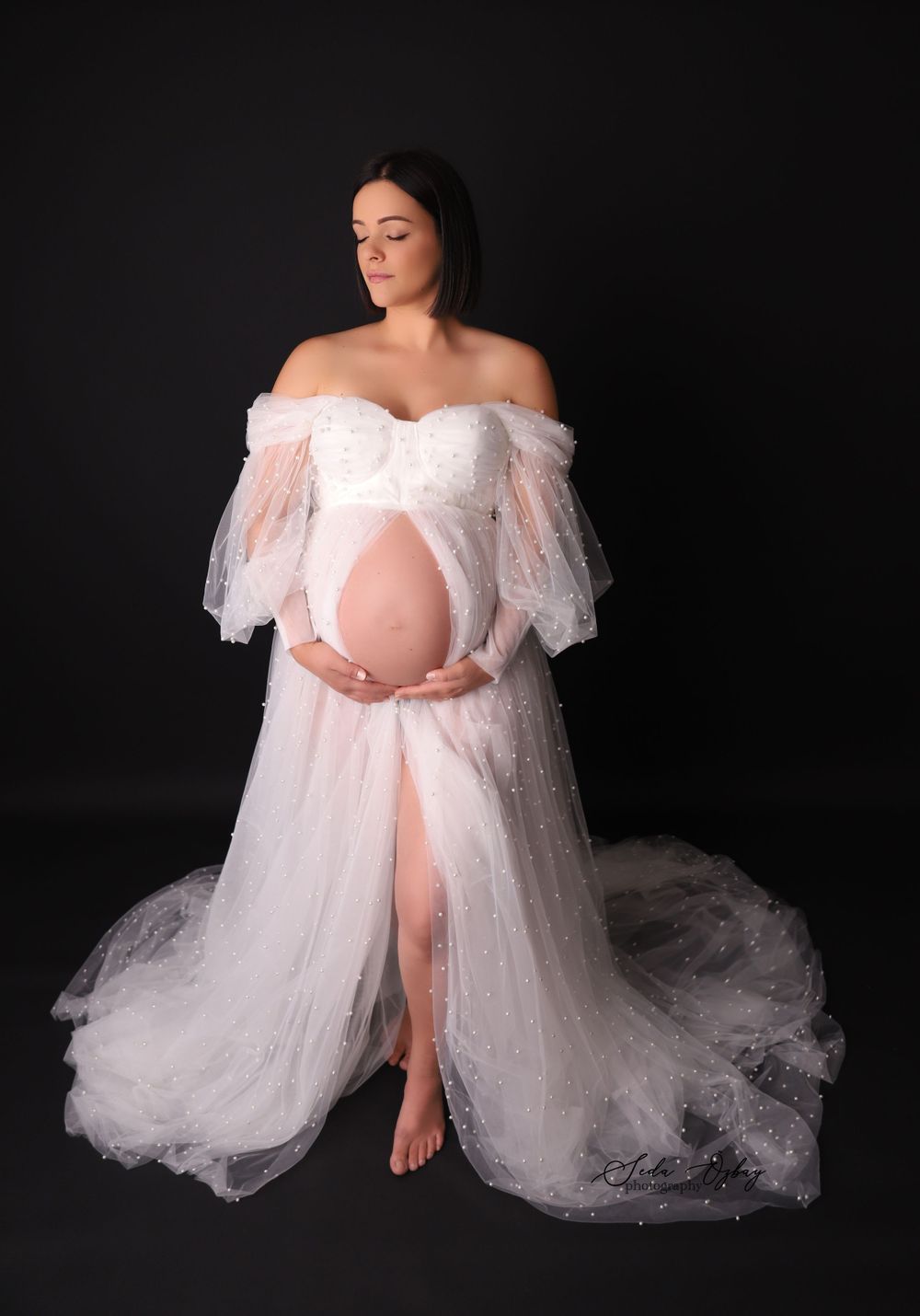 Ethereal maternity portraits in off-shoulder white lace gown against black backdrop.