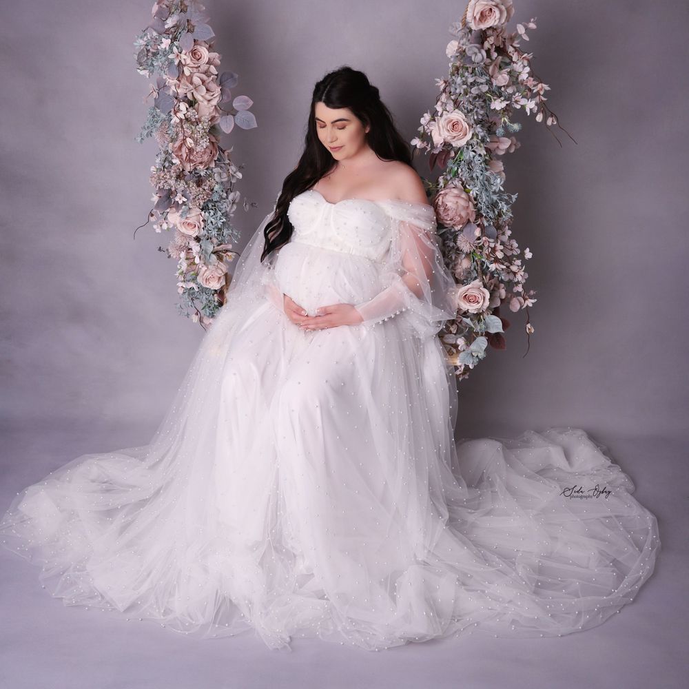 Romantic maternity portraits with floral arrangements and flowing white gown against purple backdrop.