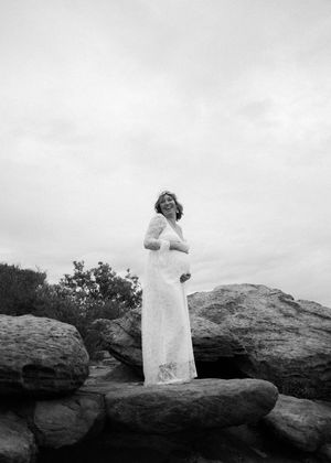 A person in a flowing white dress stands gracefully atop rocky terrain against an overcast sky.