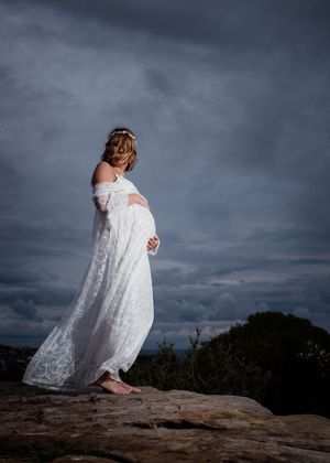 Dramatic maternity photos in white lace dress against stormy sky on rocky terrain.