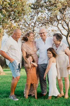 Multi-generational family gathering under trees in a sunny field wearing casual summer attire.