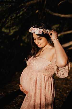 An expectant mother in a pink lace dress adjusts her floral crown in golden sunlight.