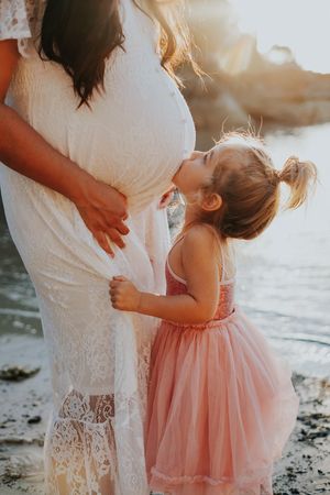 Mother and daughter in white and pink dresses share tender moment on beach at sunset.