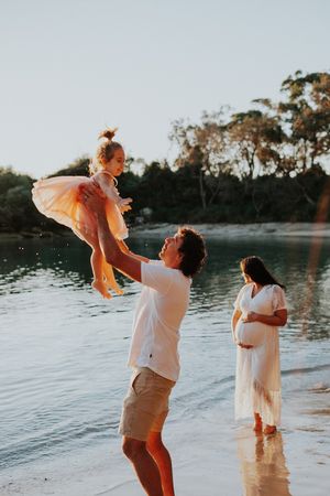 Family playing and lifting child in the air by water during golden hour.