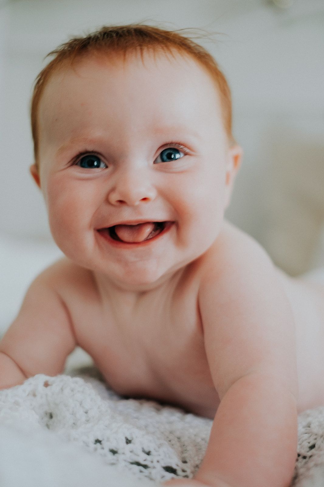 Adorable baby with bright blue eyes lies on white lace blanket with a sweet smile.