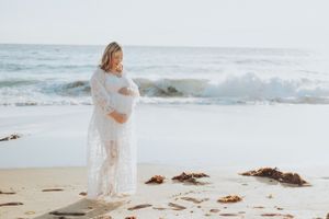 Woman in flowing white dress walks along sandy beach near ocean waves.