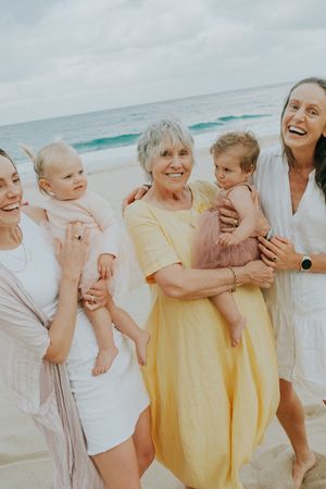A joyful beach gathering with multiple generations in white and yellow dresses against ocean waves.