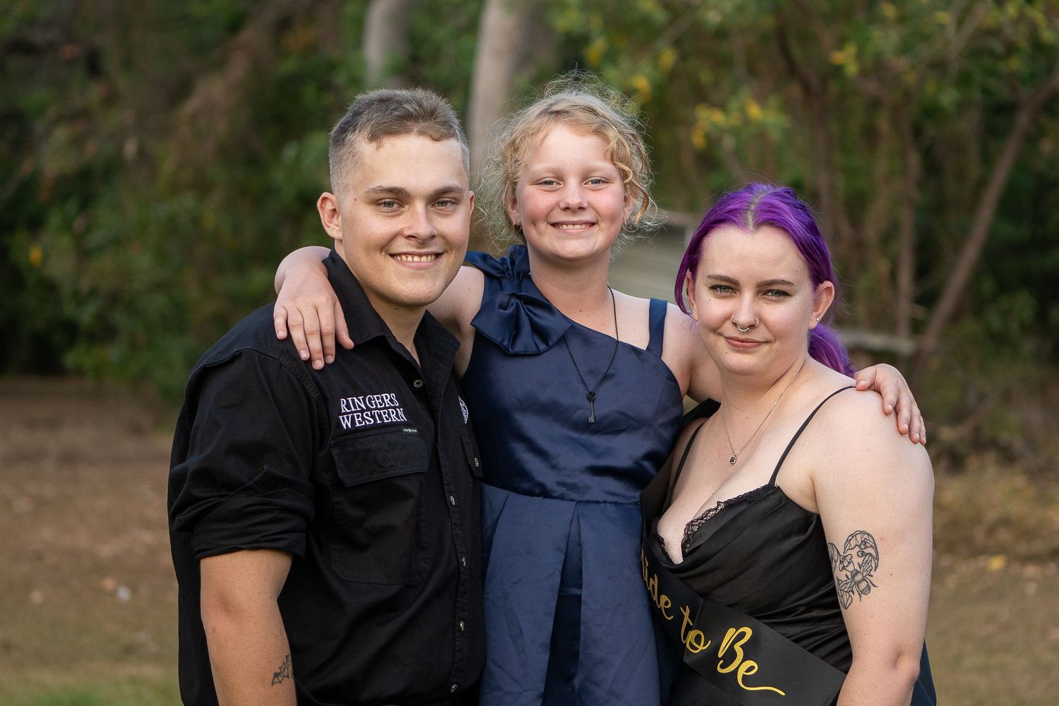 A group of people pose together outdoors in casual attire with trees in the background.