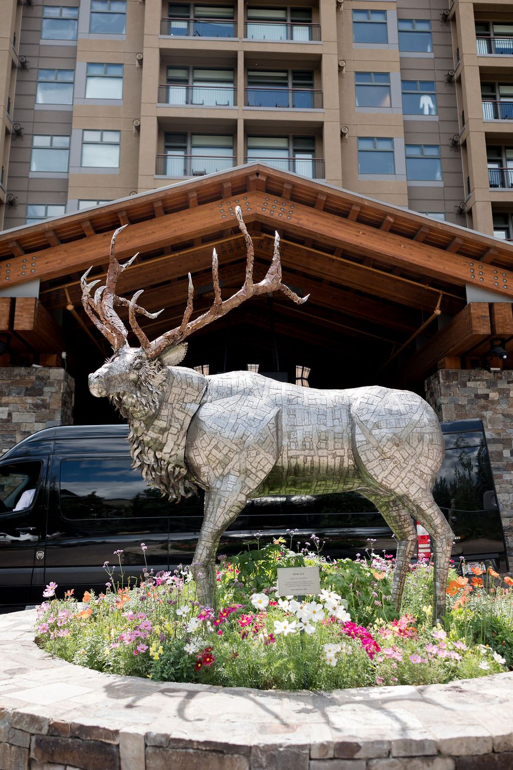 Large metal moose sculpture displayed in a garden bed with colorful flowers outside a modern building.