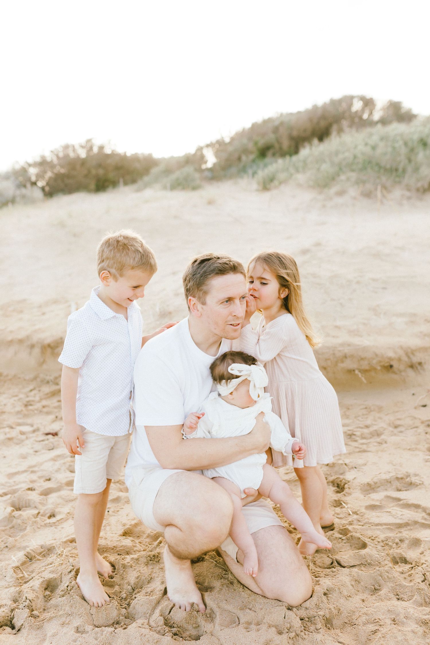 A family in white and neutral colored clothing sits together on a sandy beach during golden hour.