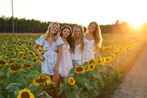 Group of friends in white dresses walking through a sunflower field at sunset.
