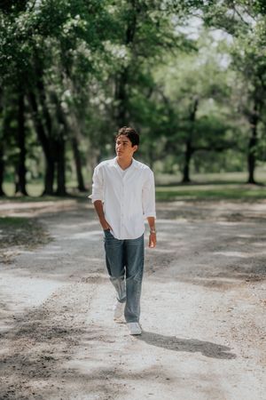 Young man in white shirt and jeans walking on a sunlit park path surrounded by lush green trees.