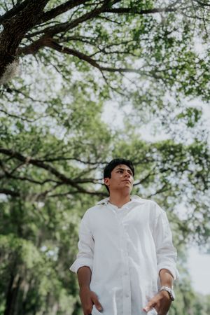 Young man in white linen shirt standing under lush green trees, looking upward in a serene outdoor setting.