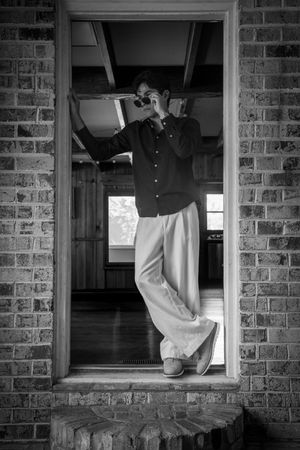 Black and white photo of a person in wide-leg pants and dark shirt posing in a brick doorway wearing sunglasses.