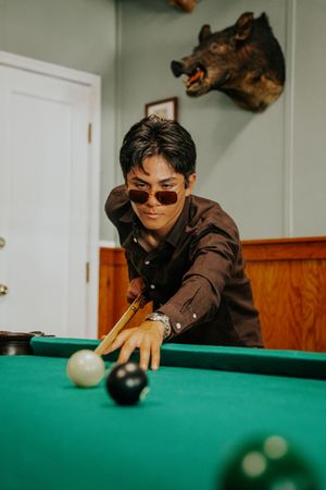 Man in sunglasses playing pool, leaning over green billiard table with cue stick, animal trophy mount on wall behind.