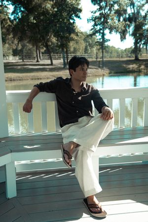 Young man in black shirt and white pants relaxing on a white porch bench near a serene pond surrounded by trees.