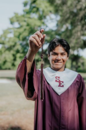 Smiling graduate in maroon SE gown holds up 2023 tassel outdoors with green trees in background.