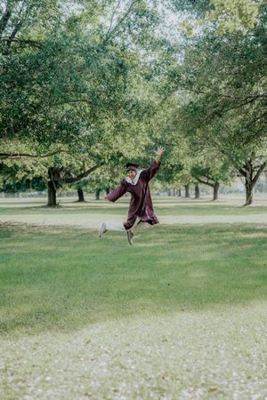 Graduate in maroon gown joyfully jumping in a lush green park, celebrating academic achievement outdoors.