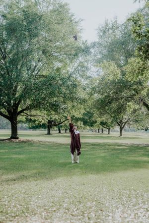 Graduate in maroon gown stands in lush green park, tossing cap in the air among tall trees.