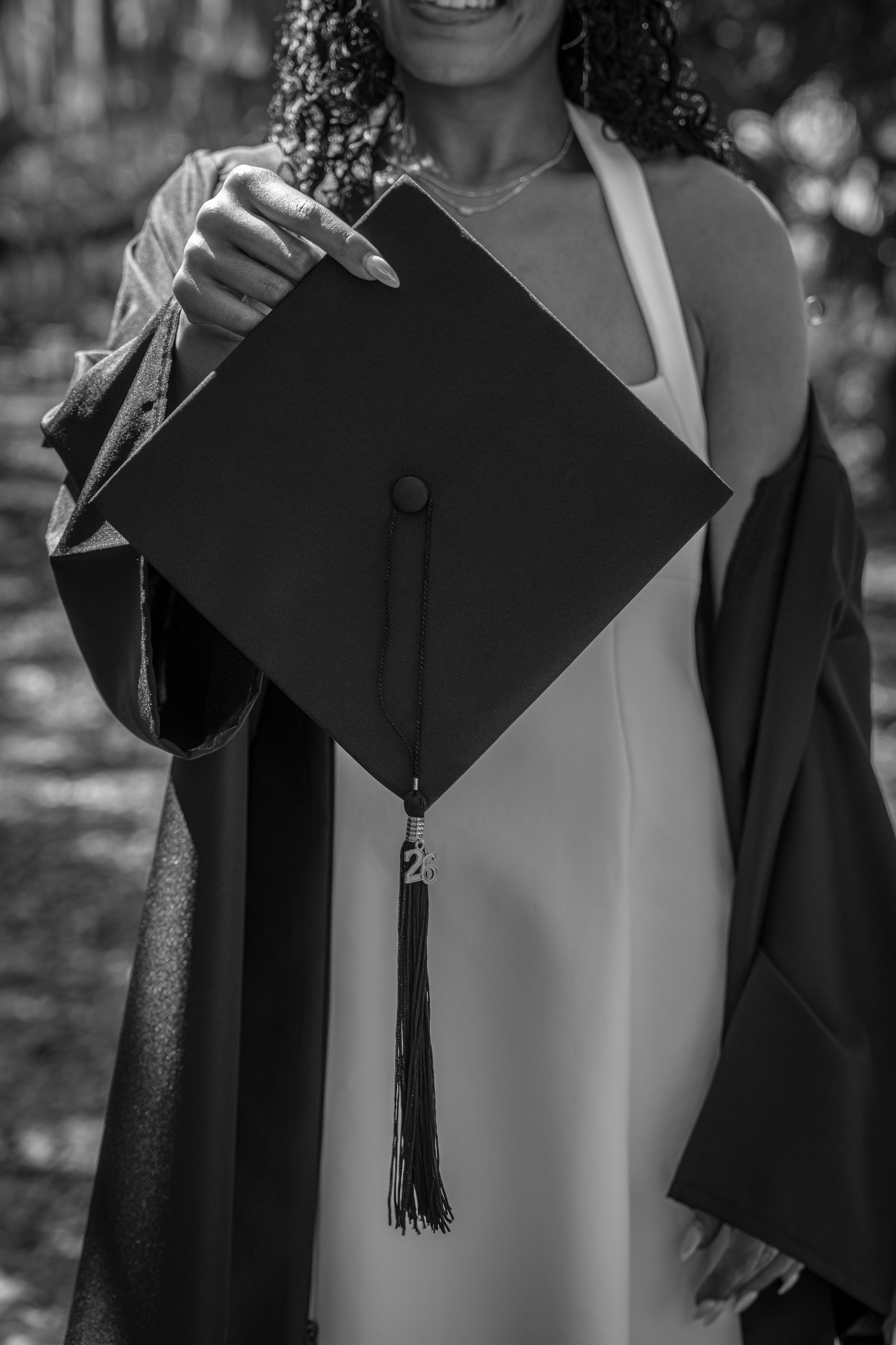 Black and white photo of a smiling graduate in cap and gown holding her mortarboard toward the camera.