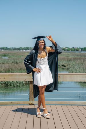 Smiling graduate in black cap and gown over white dress poses on wooden dock with marsh background.