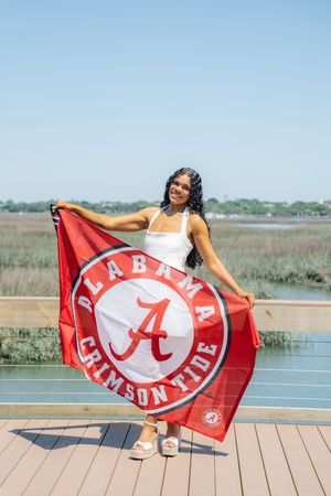 Smiling young woman proudly displaying an Alabama Crimson Tide flag on a sunny outdoor deck.