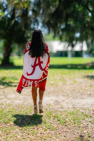 Young woman walking outdoors draped in University of Alabama Crimson Tide flag in a sunny green park.