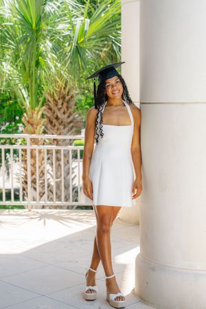 Young woman in white dress and graduation cap leaning against a column outdoors with palm trees.