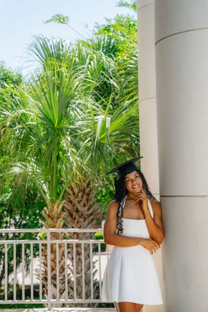 Smiling graduate in white dress and cap leaning against a column with tropical palms in background.