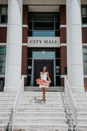 Young woman in white dress holds Bama Bound sign on City Hall steps in celebratory photo.