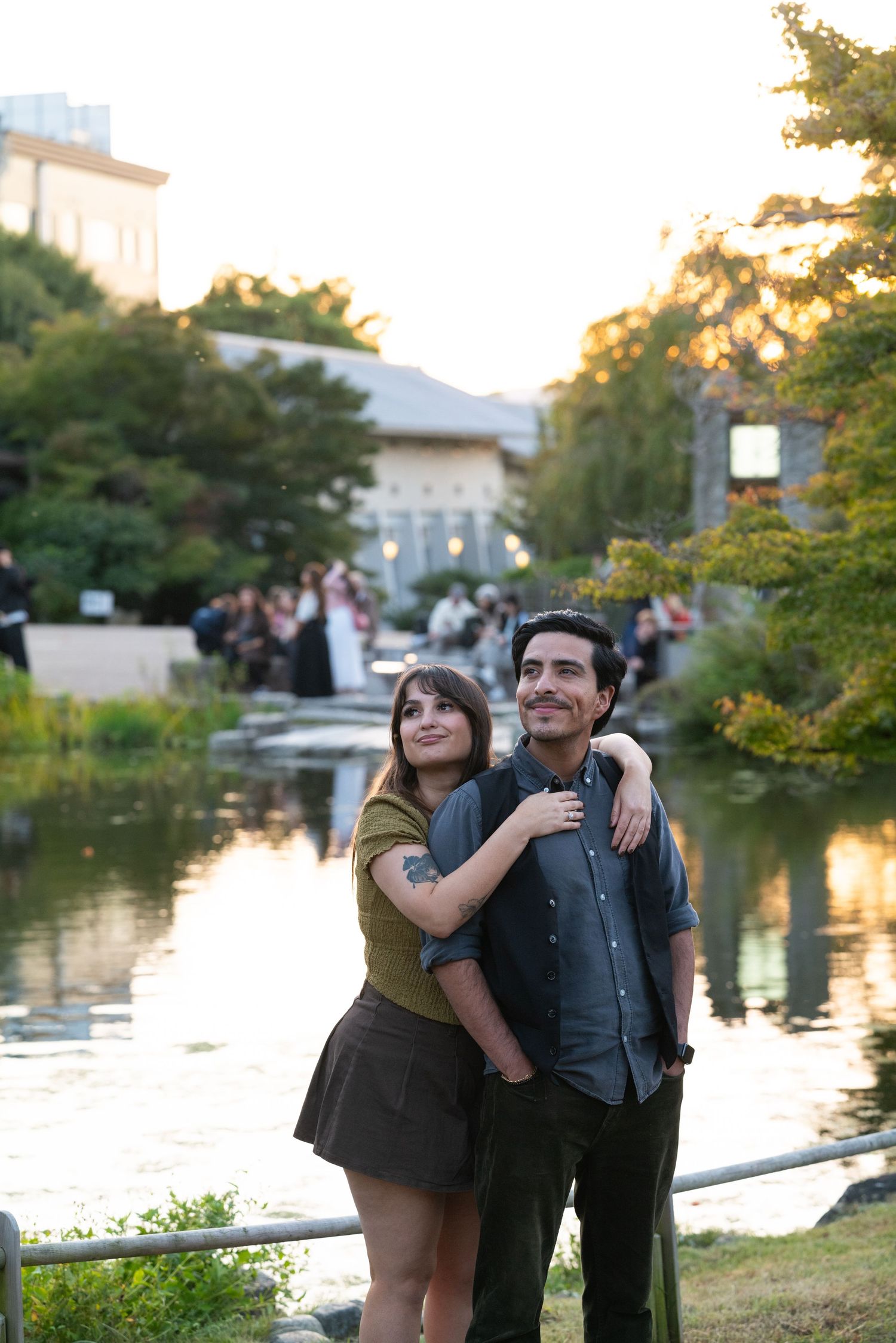 Couple embracing and gazing upward by a serene pond at sunset during their engagement photo session.